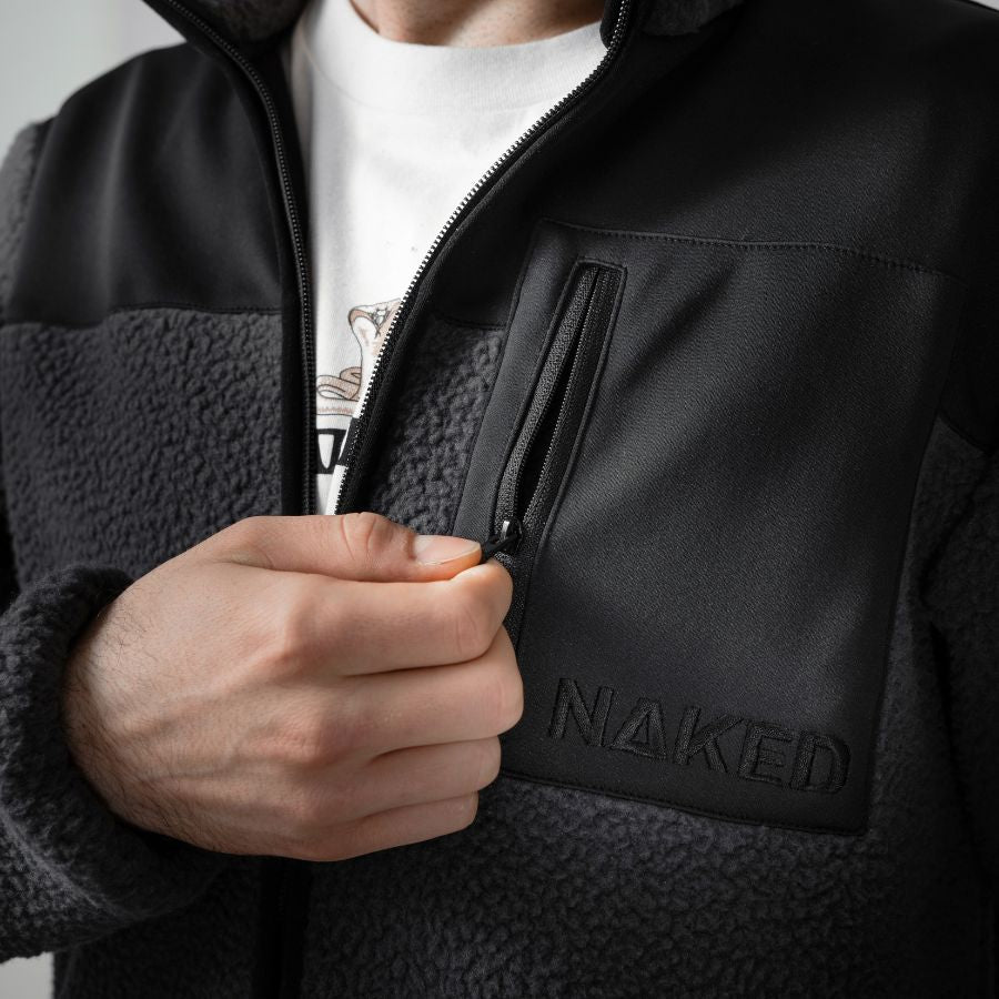 Eine Person öffnet die Brusttasche einer schwarz-grauen, winddichten Fleecejacke von NAKED Optics mit dem NAKED-Logo auf der Tasche.