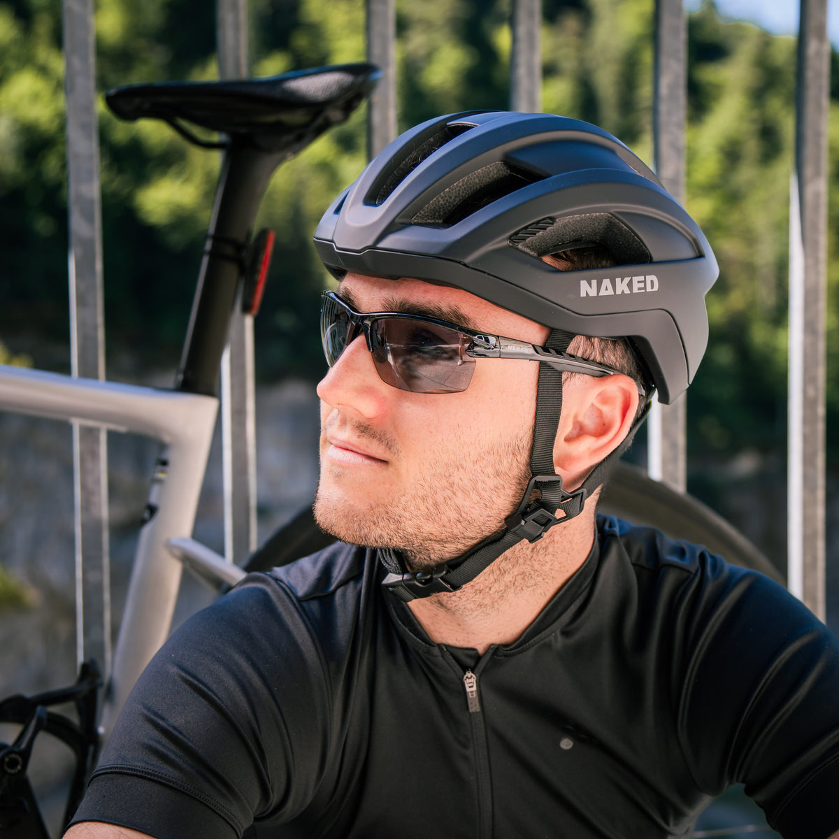 Ein Mann in Radbekleidung und mit Helm, der die „The RUSH Halfframe“ von NAKED Optics trägt – eine photochrome Sportbrille mit selbsttönenden Gläsern für optimale Sicht und UV400-Schutz –, sitzt neben seinem Fahrrad und blickt vor einem verschwommenen Außenhintergrund nach oben.
