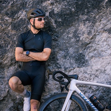 Ein Radfahrer in schwarzer Fahrbekleidung und mit Helm steht mit verschränkten Armen an einer Felswand neben seinem Rennrad – sein Look wird durch die elegante RUSH Halfframe Photochromisch von NAKED Optics abgerundet, die mit UV400-Schutz und selbsttönenden Gläsern für ultimativen Augenschutz sorgt.