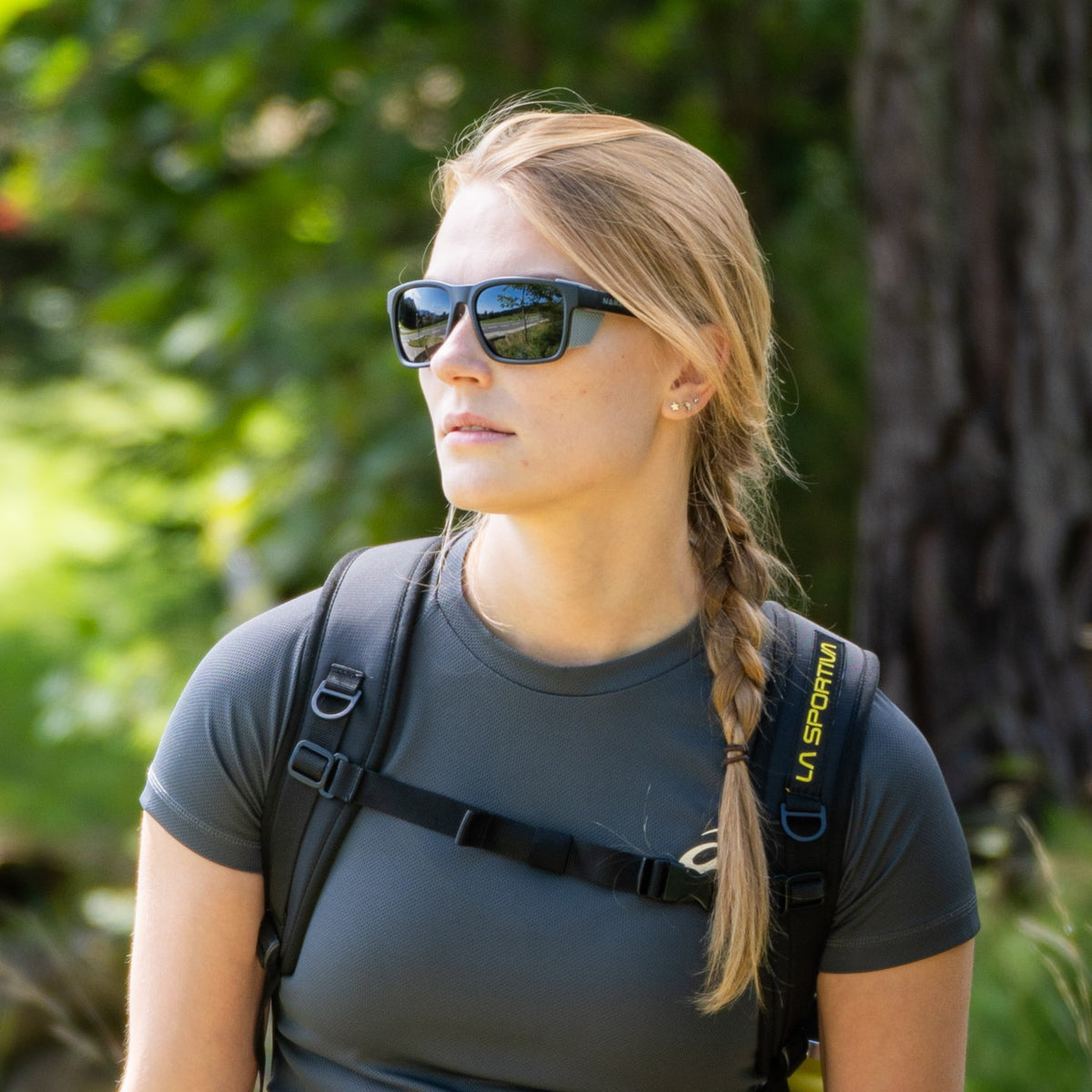 A woman with sunglasses and a braid, wearing a backpack and standing outdoors surrounded by greenery, is ready for alpine Abenteuer with her stylish “The ASCENT” Gletscherbrille from NAKED Optics, offering UV400 Schutz.