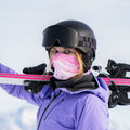 Eine Person in lila Jacke und Helm trägt rosa Skier auf der Schulter und hat die „The SOLAR“ – eine kompakte Damen-Skibrille von NAKED Optics – sowie eine Skimaske und eine Schutzbrille aufgesetzt; im Hintergrund ist Schnee zu sehen.