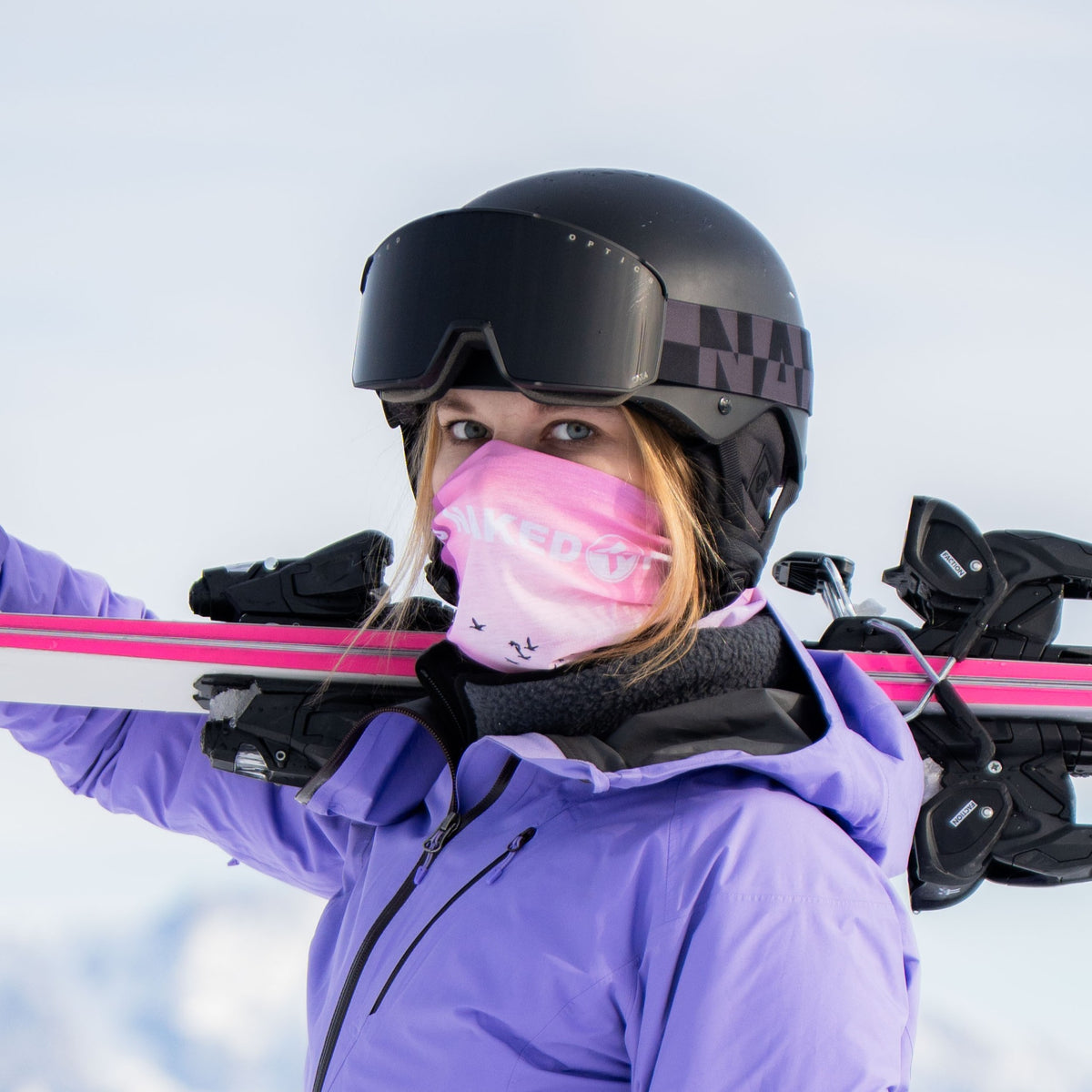 Eine Person in lila Jacke und Helm trägt rosa Skier auf der Schulter und hat die „The SOLAR“ – eine kompakte Damen-Skibrille von NAKED Optics – sowie eine Skimaske und eine Schutzbrille aufgesetzt; im Hintergrund ist Schnee zu sehen.