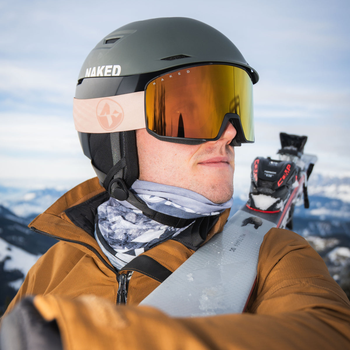Un uomo in tenuta da sci e casco, che indossa gli occhiali da sci The SOLAR kompakte di NAKED Optics, tiene gli sci in spalla con le montagne innevate sullo sfondo.