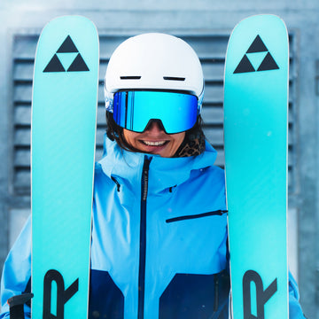 A person in blue ski gear and helmet, smiling while holding blue skis, wears the NAKED Optics The SOLAR—reflektive Skibrille Damen und kompakte Skibrille für kleine Gesichter—in front of a snowy background.