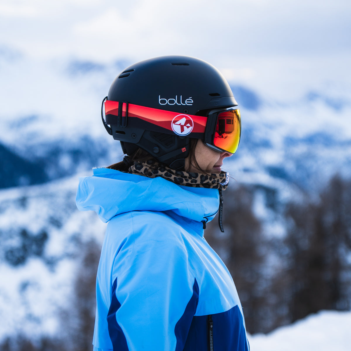A person in a blue ski jacket and helmet, wearing NAKED Optics’ The STORM skibrille sphärisch with Anti-Fog-Beschichtung, stands sideways in a snowy mountain landscape.