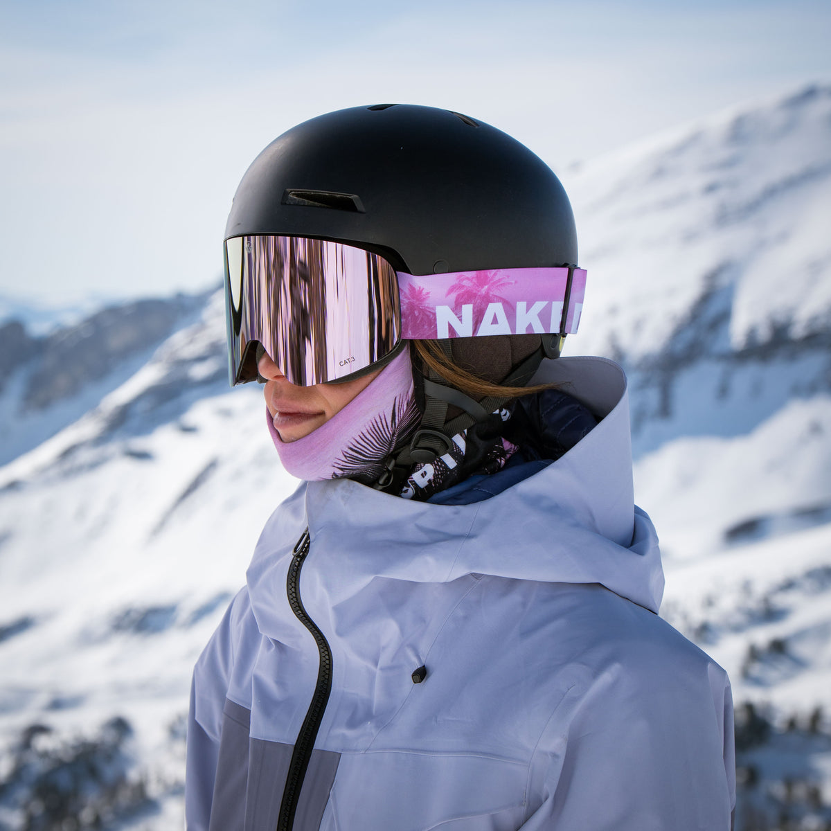 A person in ski gear and a helmet stands in snowy mountains, wearing The TROOP EVO pink ski goggles from NAKED Optics with anti-fog coating and a white jacket.