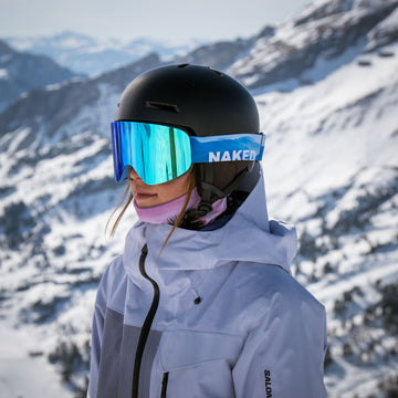 Persona in tenuta da sci con gli occhiali da sci blu “TROOP EVO” di NAKED Optics, dotati di rivestimento antiappannamento, e casco nero, in piedi su un pendio innevato.