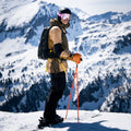 A skier in tan and black gear, wearing the TROOP EVO ski goggles from NAKED Optics with anti-fog coating, stands on a snowy mountain with poles, snowy peaks and a blue sky in the background.