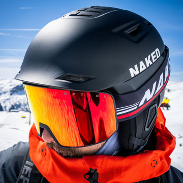 Ein Wintersportler trägt einen schwarzen APEX-Helm von NAKED Optics und eine orangefarbene Spiegelbrille.
