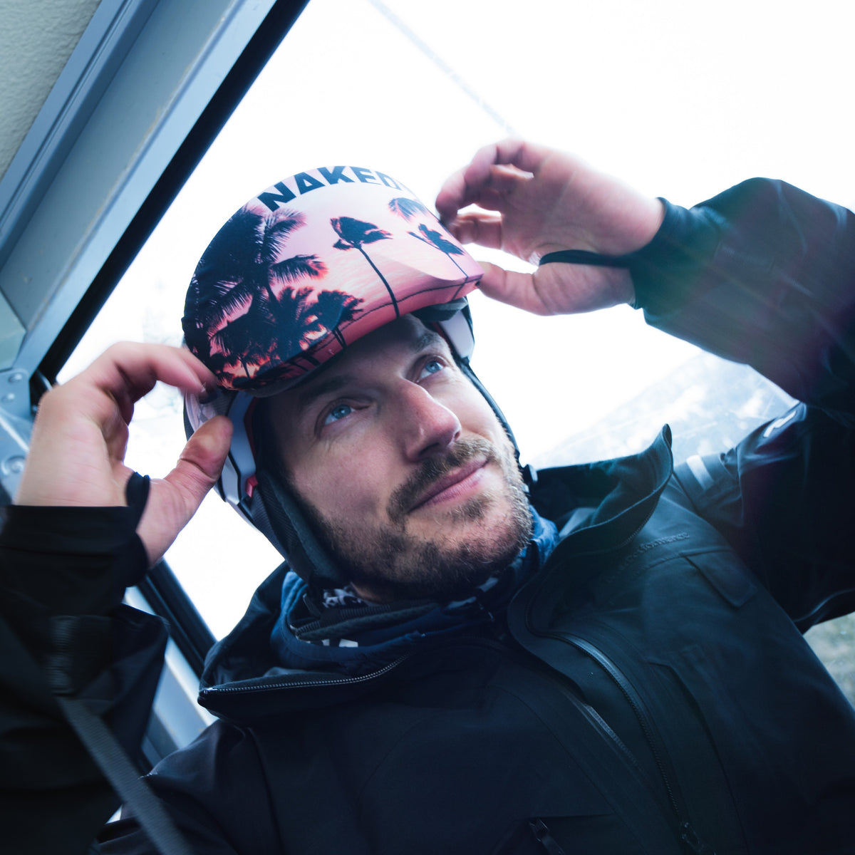 Un uomo si sistema un casco rosa con un disegno di una palma, indossando una giacca all'interno di un veicolo con i finestrini chiusi, mentre i suoi NAKED Optics Skibrillen Schutz (Überzieher) proteggono i suoi occhiali da sci conferendo loro un tocco di stile.