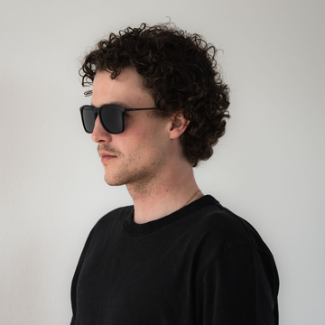 Een man met krullend haar, die de zwarte ACE-zonnebril met gepolariseerde glazen van NAKED Optics draagt en een zwart shirt, staande tegen een effen witte achtergrond.