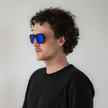 A man with curly hair wearing The ACE Sonnenbrille by NAKED Optics, featuring black frames and blue reflective, polarisierte Gläser, stands against a plain white background, ensuring both UV-Schutz and modern style.
