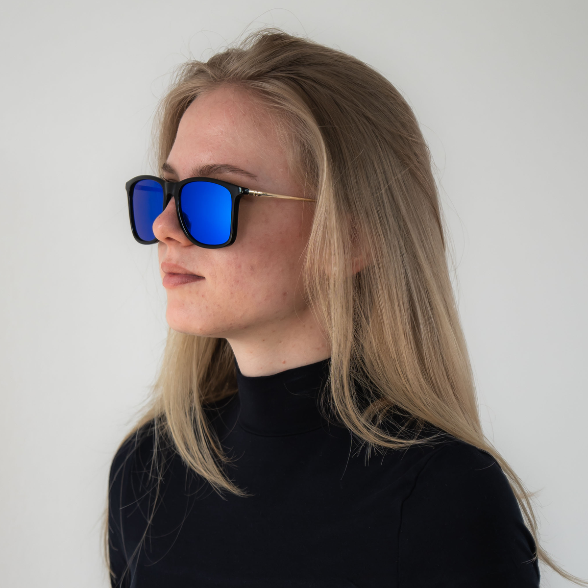 Woman with long blonde hair in a black turtleneck, wearing NAKED Optics “The ACE” reflective blue sunglasses with UV protection, looks left.