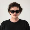 Ein Mann mit lockigem Haar trägt die schwarze Sonnenbrille The AURA von NAKED Optics im Retro-Design und ein schwarzes T-Shirt, während er vor einem schlichten Hintergrund steht.