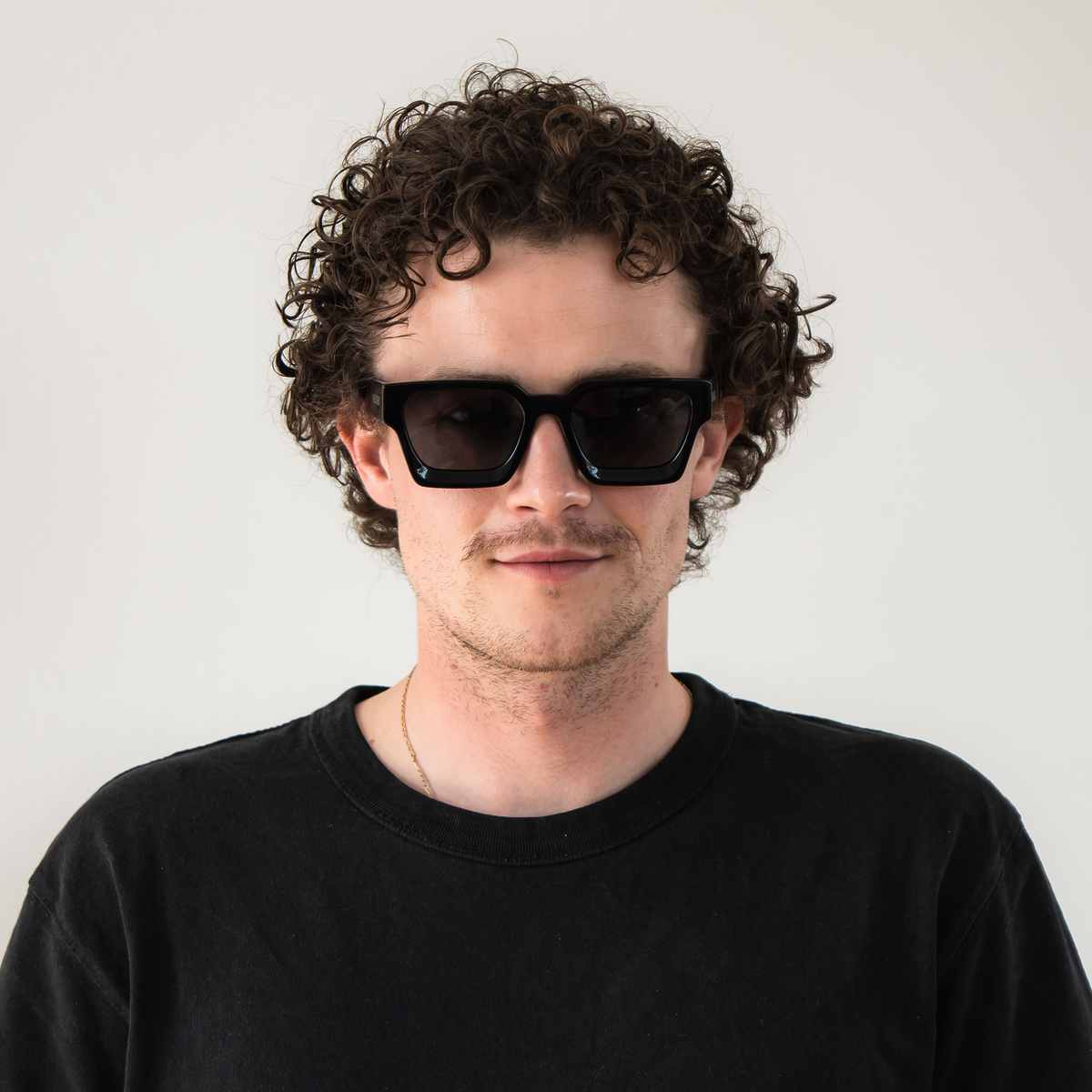 Ein Mann mit lockigem Haar trägt die schwarze Sonnenbrille The AURA von NAKED Optics im Retro-Design und ein schwarzes T-Shirt, während er vor einem schlichten Hintergrund steht.