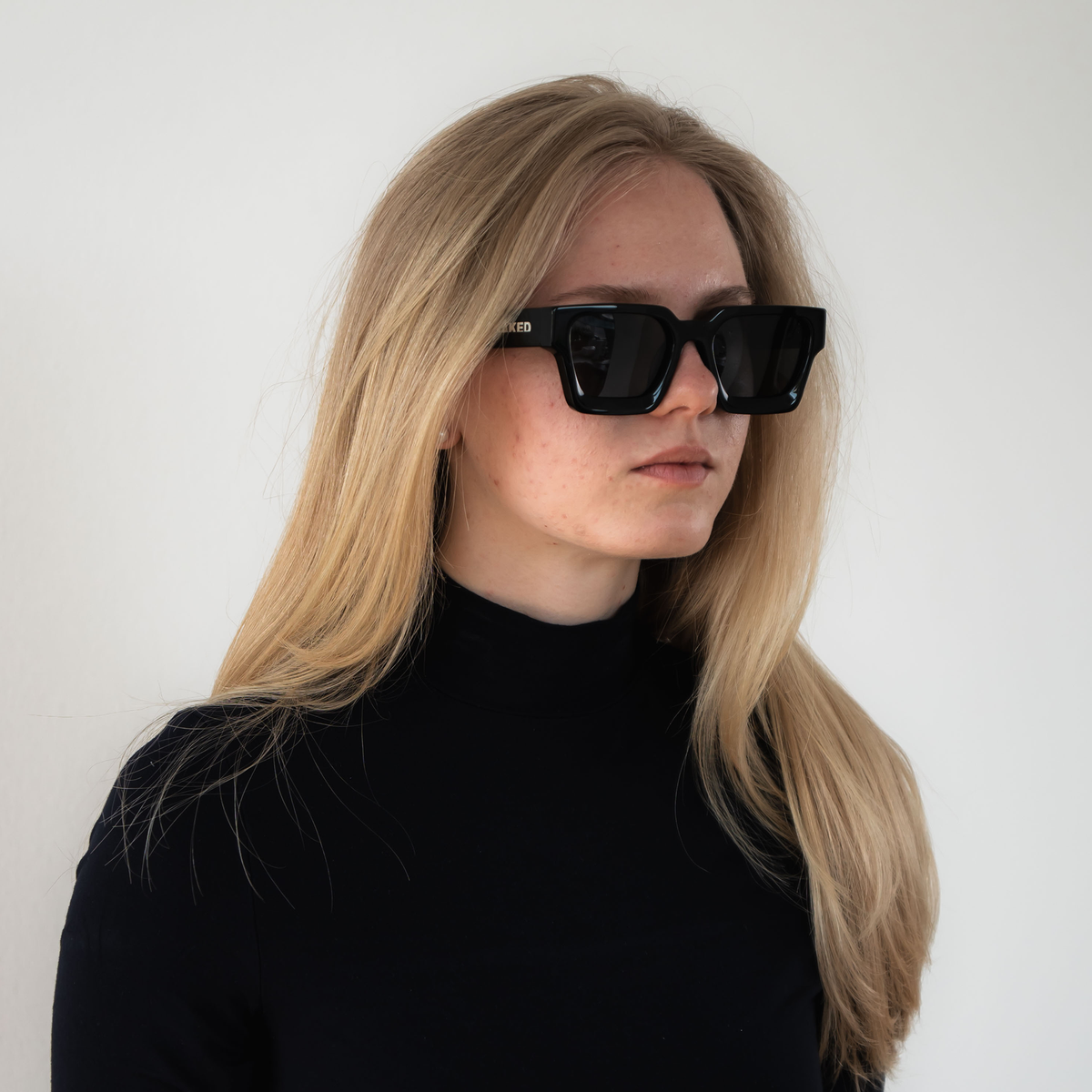 A woman with long blonde hair wears The AURA black sunglasses with polarisierte Gläser by NAKED Optics, styled with a black turtleneck against a plain background.