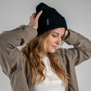 Een vrouw met lang haar draagt een gezellige NAKED Optics Beanie BASE en een jasje; ze kijkt naar beneden en glimlacht tegen een neutrale achtergrond.