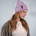 Eine Frau mit gewelltem Haar trägt eine lavendelfarbene Beanie „BASE“ von NAKED Optics aus weichem Acryl sowie eine taupefarbene Jacke und blickt vor einem schlichten Hintergrund nach unten.