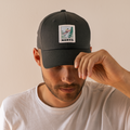 Un uomo in camicia bianca si sistema il cappellino da baseball scuro della NAKED Optics, un modello unisex realizzato in cotone di alta qualità e caratterizzato da una toppa con la scritta “NAKED” e un’illustrazione che raffigura un surfista.