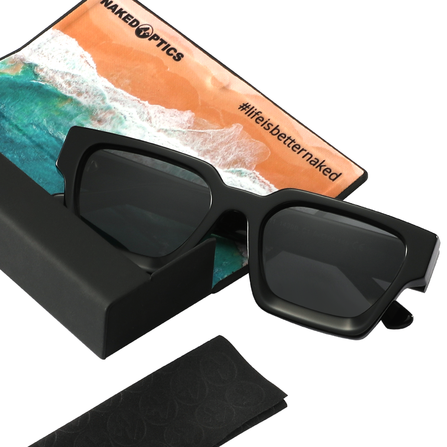 Die schwarze Sonnenbrille The AURA von NAKED Optics mit polarisierten Gläsern liegt neben einem Reinigungstuch mit Strandmotiv und der Aufschrift #lifeisbetternaked – das perfekte Accessoire im angesagten Retro-Design.