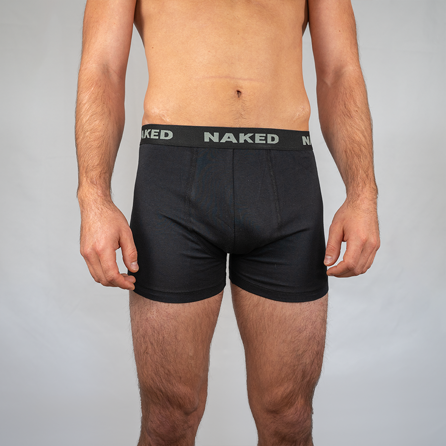 Ein Mann trägt schwarze Boxershorts – 2er-Pack von NAKED Optics – mit einem Bund, der das NAKED-Logo trägt, während er vor einem schlichten hellgrauen Hintergrund steht. Diese Boxershorts aus Baumwolle von NAKED Optics bieten außergewöhnlich atmungsaktive Unterwäsche und optimalen Tragekomfort für den Alltag.