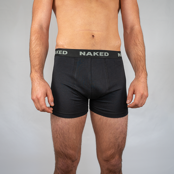 Ein Mann trägt schwarze Boxershorts – 2er-Pack von NAKED Optics – mit einem Bund, der das NAKED-Logo trägt, während er vor einem schlichten hellgrauen Hintergrund steht. Diese Boxershorts aus Baumwolle von NAKED Optics bieten außergewöhnlich atmungsaktive Unterwäsche und optimalen Tragekomfort für den Alltag.