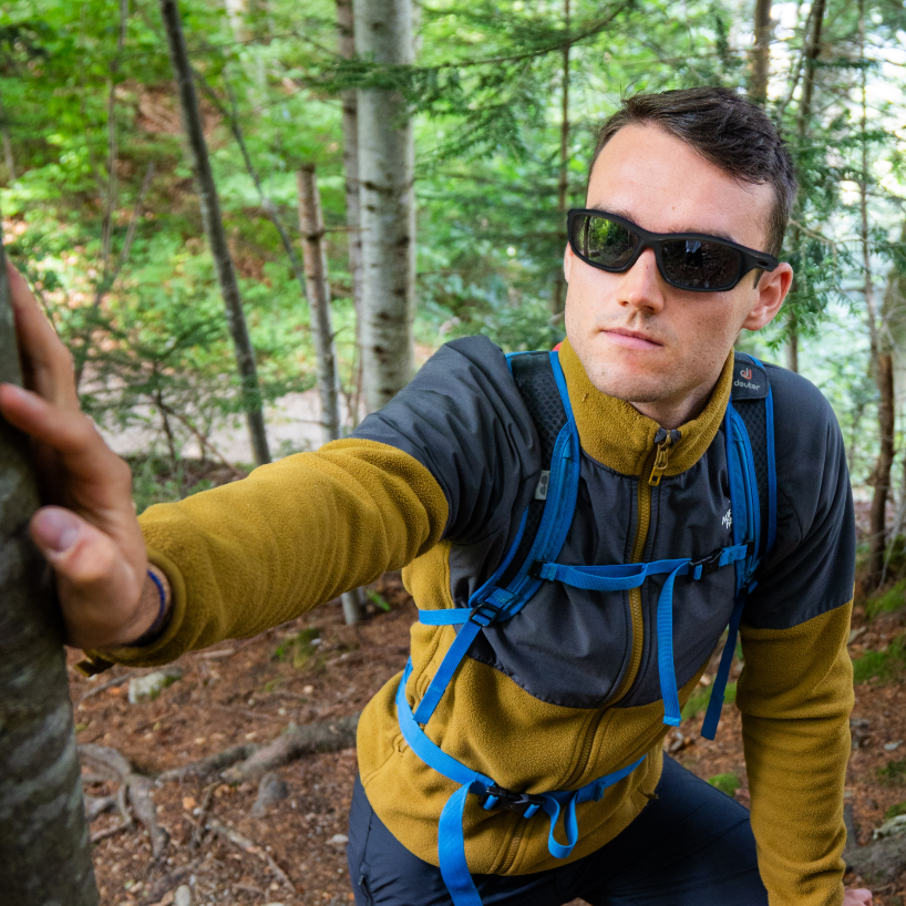 Un randonneur portant des lunettes de soleil The RAVE de la marque NAKED Optics et un sac à dos est adossé à un arbre de la forêt.