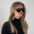 Woman with long blonde hair and sunglasses wearing a black turtleneck, facing left against a white wall. She completes her look with The CARTEL by NAKED Optics, adding a touch of vintage flair to her modern style.