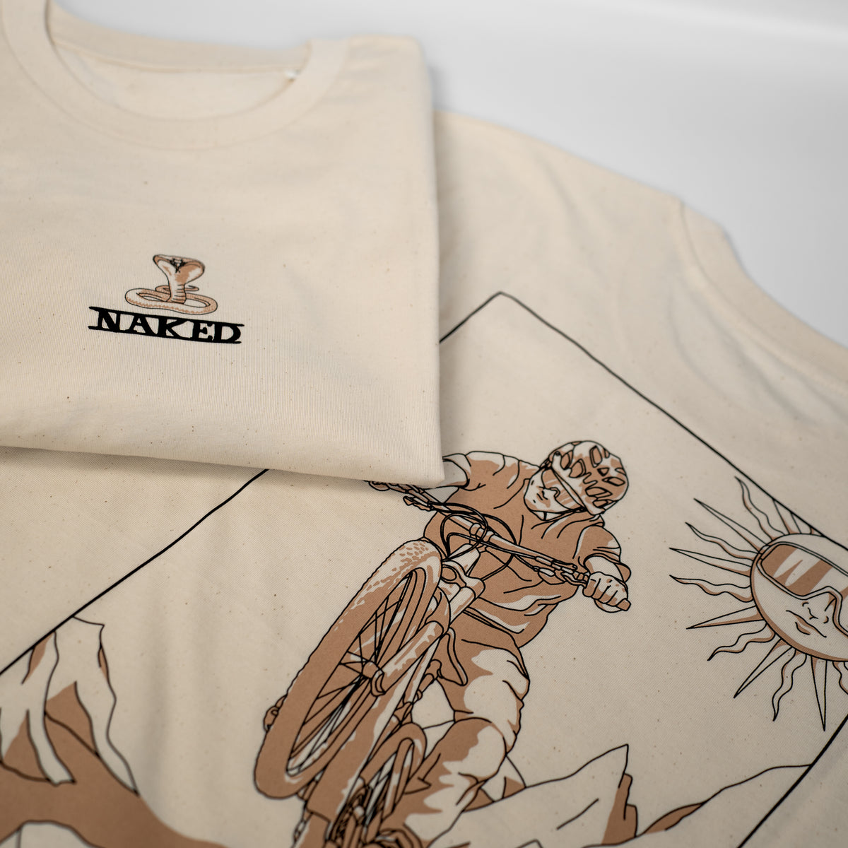 Due magliette unisex beige di NAKED Optics: una con il logo del serpente “The Fool” e l’altra con l’illustrazione di un ciclista di mountain bike e del sole ispirata a Stevie Schneider — una maglietta imperdibile per gli appassionati di ciclismo.