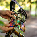 Mani che reggono due lenti intercambiabili Ersatzglas ZEND di NAKED Optics — una trasparente e una iridescente — dotate di un semplice sistema di sostituzione delle lenti a magneti (Magnetsystem Gläserwechsel) per cambi rapidi e una protezione UV-400 affidabile, ritratte accanto a una mountain bike.