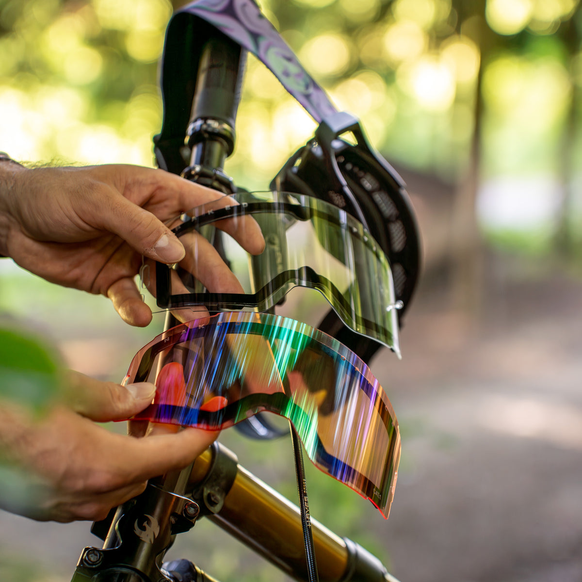 Mani che reggono due lenti intercambiabili Ersatzglas ZEND di NAKED Optics — una trasparente e una iridescente — dotate di un semplice sistema di sostituzione delle lenti a magneti (Magnetsystem Gläserwechsel) per cambi rapidi e una protezione UV-400 affidabile, ritratte accanto a una mountain bike.