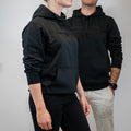 Two people in dark hoodies stand side by side, facing slightly away from the camera against a plain background. Each wears a NAKED Optics Hoodie Signature Stitch in black with a subtle embroidered logo, making these pullovers both stylish and versatile.