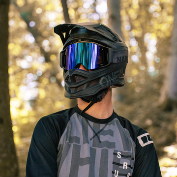 Una persona con un casco da bicicletta nero e occhiali protettivi, che indossa una maglia grigia e sfoggia gli occhiali da MTB di alta qualità “The ZEND” di NAKED Optics, si trova in un bosco illuminato dal sole.