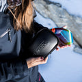 Eine Person hält in einer verschneiten Landschaft im Freien eine Skibrille mit Regenbogenglas, die mit dem innovativen SnapTech-Magnetwechselsystem ausgestattet ist, sowie ein schwarzes NAKED Optics-Etui für Wechselgläser in der Hand.