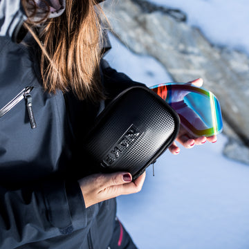 A person holds Skibrillen with a rainbow lens featuring the innovative SnapTech-Magnetwechselsystem and a NAKED Optics Etui für Wechselgläser in black in a snowy outdoor setting.