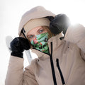 A person in a beige jacket and beanie, wearing black gloves and using a colorful NAKED Optics Schlauchschal (Bandana) as a face covering, standing in the snow.