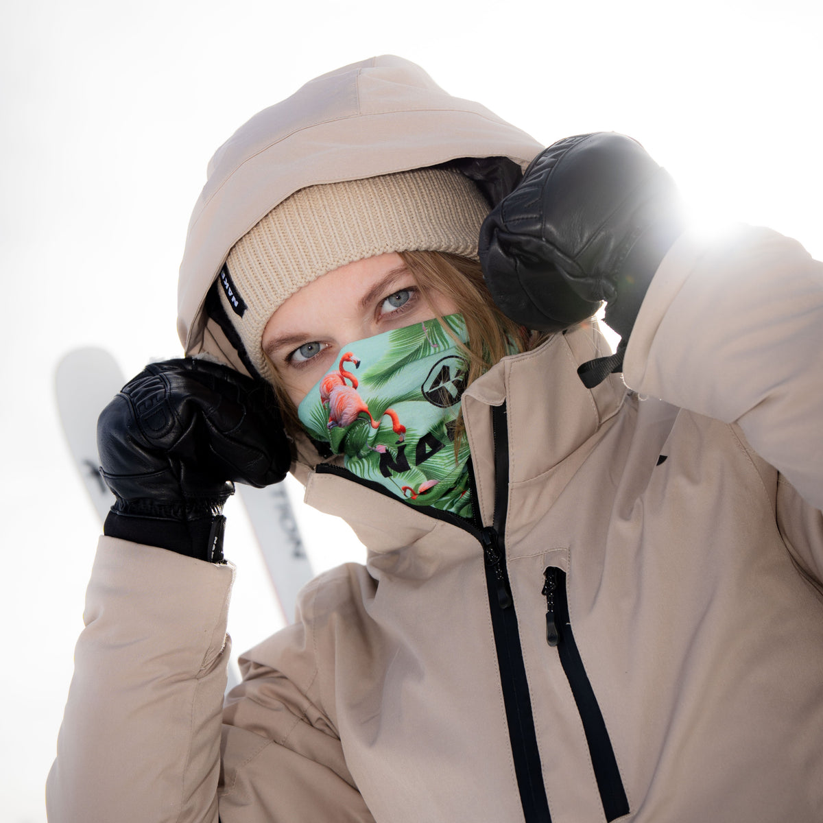 A person in a beige jacket and beanie, wearing black gloves and using a colorful NAKED Optics Schlauchschal (Bandana) as a face covering, standing in the snow.