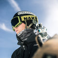 A skier in goggles and helmet looks ahead, holding skis over their shoulder against a bright blue sky, showcasing a NAKED Optics Schlauchschal (Bandana) that adds both comfort and style to their winter gear.