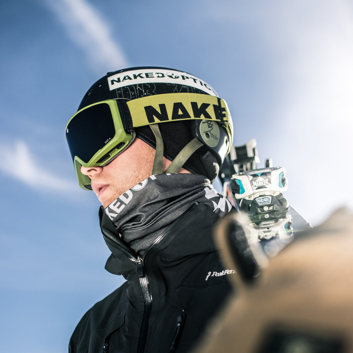 A skier in goggles and helmet looks ahead, holding skis over their shoulder against a bright blue sky, showcasing a NAKED Optics Schlauchschal (Bandana) that adds both comfort and style to their winter gear.