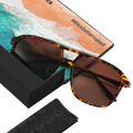 Tortoiseshell sunglasses with brown lenses rest on a box, accompanied by a beach postcard and the visible hashtag #lifeisbetternaked—these “The CARTEL” sunglasses from NAKED Optics feature a classic retro design, making them perfect for sunny days.