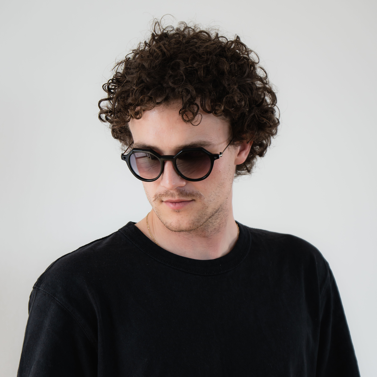 Man with curly hair wearing The IZZY sunglasses by NAKED Optics, featuring an acetate frame and a black shirt, looking slightly down and to the side.