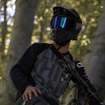 Con indosso un casco e gli occhiali The LINE di NAKED Optics, una persona è seduta su una mountain bike in un bosco illuminato dal sole e guarda di lato.