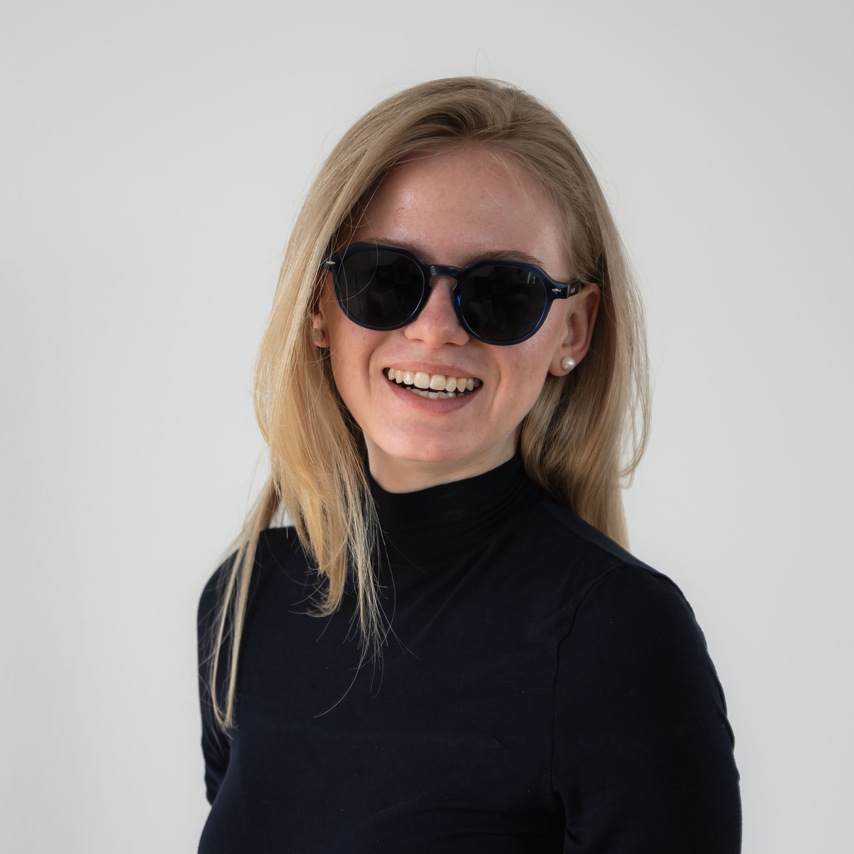 Smiling woman with long blonde hair, wearing stylish NAKED Optics The LUNA sunglasses with polarized lenses and a black turtleneck, against a white background.