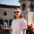 A man with curly hair wears The LUX sunglasses by NAKED Optics as he stands outside near red flowers and a statue, soaking up the sun on a beautiful day.