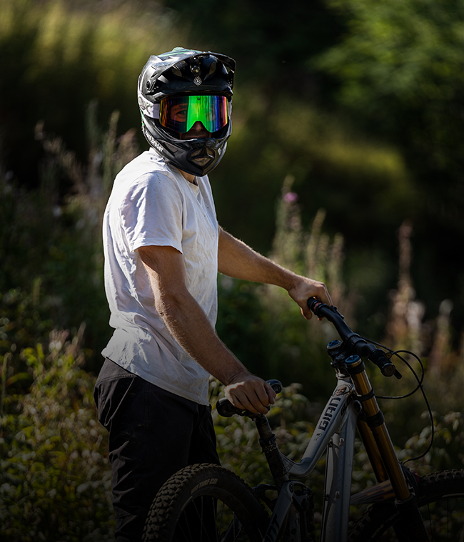 Eine Person mit Helm und Sonnenbrille steht im Freien mit einem Mountainbike, umgeben von viel Grün.