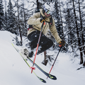Eine Person, die einen verschneiten Hang hinunterfährt und dabei abhebt, trägt einen Helm, eine beige Jacke und „The SOLAR“ von NAKED Optics – eine kompakte Skibrille, die speziell für schmale Gesichter entwickelt wurde –, im Hintergrund sind verschneite Bäume zu sehen.