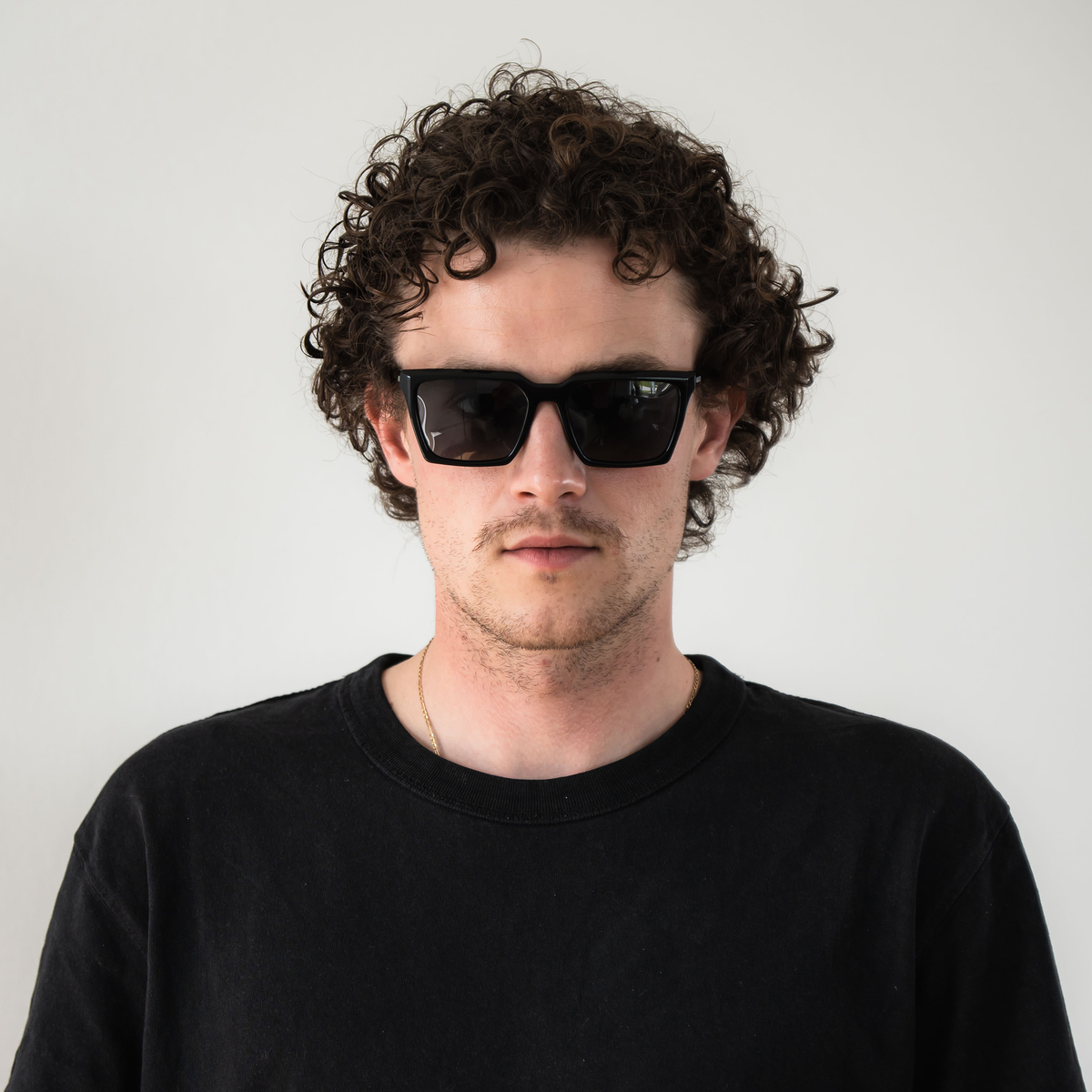 A man with curly hair wearing a black shirt and stylish NAKED Optics “The MUSE” Unisex Sonnenbrille with polarisierten Gläsern stands against a plain white background. The sunglasses provide UV400 Schutz, ensuring both fashion and eye protection.