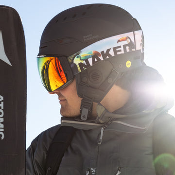 Una persona in tenuta da sci e con il casco indossa gli occhiali da sci The STORM di NAKED Optics — sferici con rivestimento antiappannamento — mentre tiene in mano gli sci ed è in piedi all’aperto con la luce del sole che splende alle sue spalle.