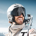 A smiling skier in a helmet and The NOVA ski goggles by NAKED Optics—with anti-fog coating—carries skis on their shoulder, snowy mountains in the background.