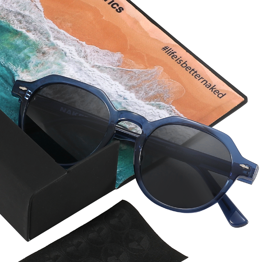 The LUNA sunglasses by NAKED Optics in blue with polarized lenses, displayed alongside a black case, cleaning cloth, and a beach-themed card that reads #lifeisbetternaked.